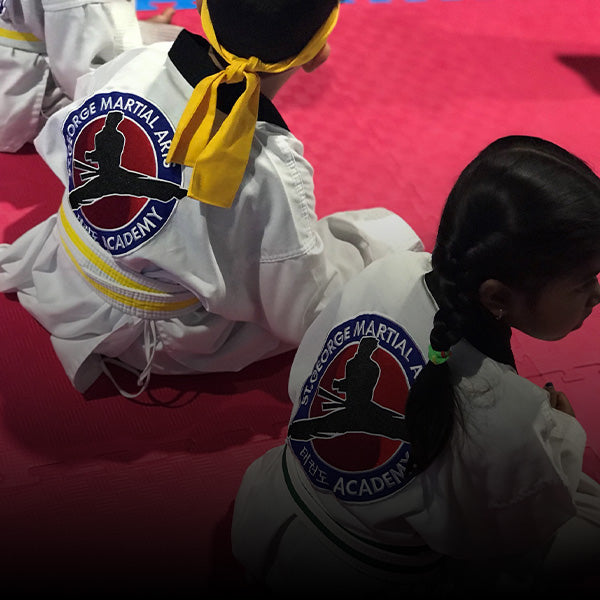 Two children wearing martial arts uniforms with "St. George Martial Arts Academy" patches sit on a pink mat, facing away from the camera. One has a yellow belt and a yellow headband; the other has braided hair.