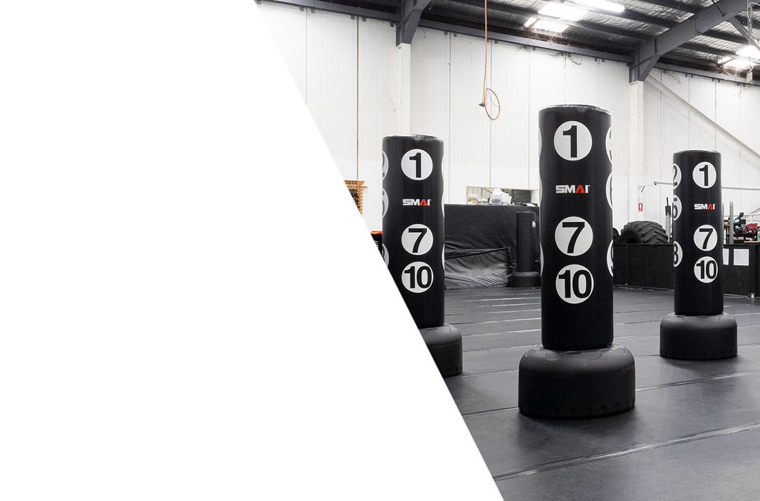 Four standing black punching bags with white numbered targets are placed in a spacious gym with padded floors and a high ceiling; the left side of the image has a large white diagonal overlay.