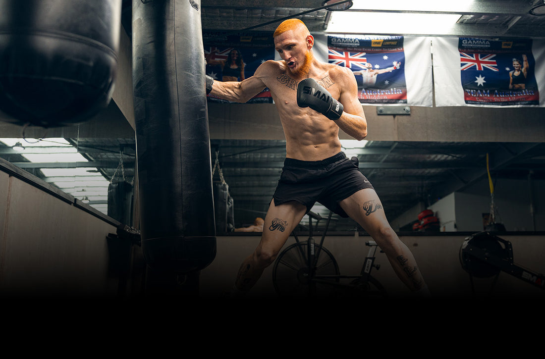 A shirtless man with orange hair wearing black boxing gloves and shorts punches a heavy bag in a gym, with flags hanging on the wall behind him.