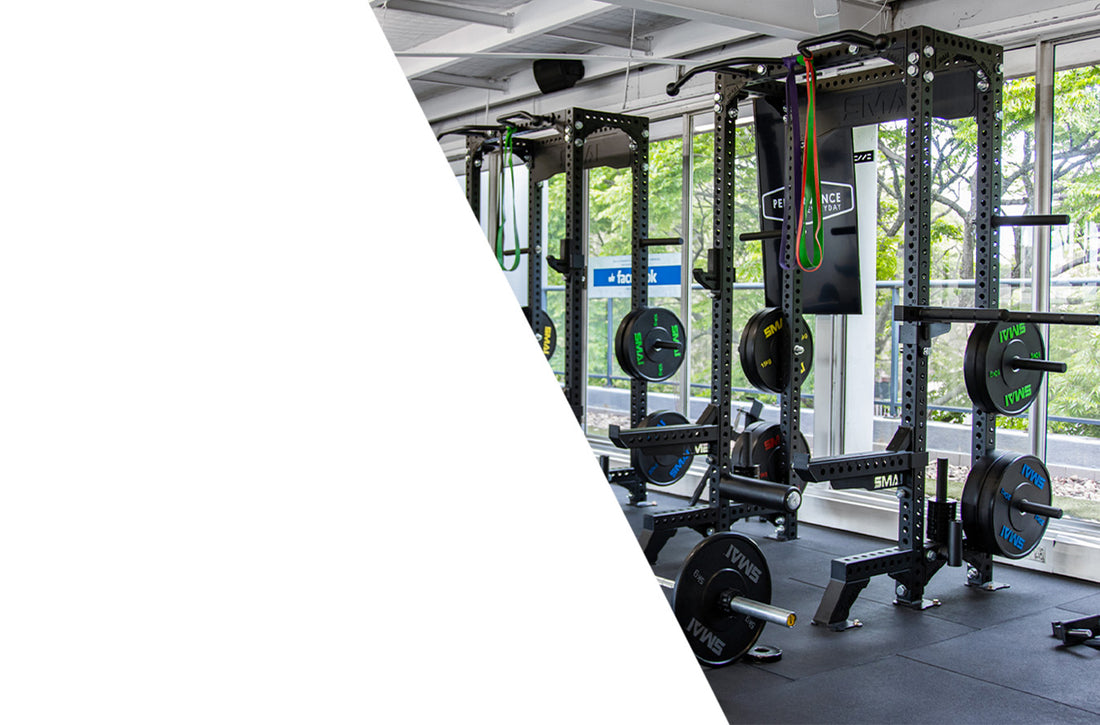 A gym with large windows, weightlifting racks, barbells, weight plates, resistance bands, and benches. Natural light fills the space, and trees are visible outside. The left side of the image is blank and white.