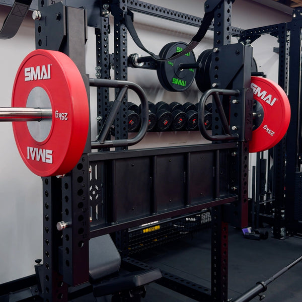 A Vanta Series Leg Press Attachment by SMAI is set up in a home gym, with red 25kg SMAI plates on the barbell, extra weights stored on the rack, and multiple dumbbells visible in the background.