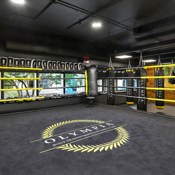 A spacious boxing gym includes a 5m SMAI Competition Boxing Ring with yellow ropes and black floor, featuring "OLYMPIA" and a laurel wreath on the mat. Equipped with commercial-grade boxing gear, punching bags are around, while large windows offer tree views.