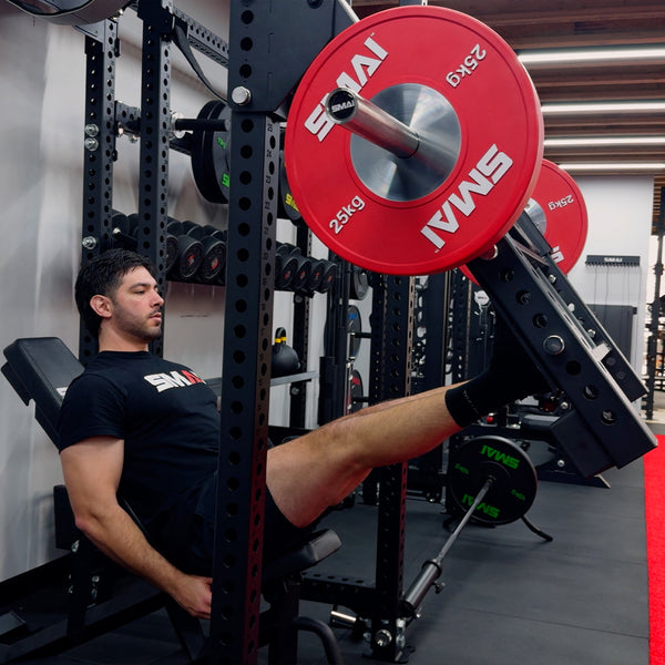 A man wearing a black T-shirt, shorts, and socks uses the SMAI Vanta Series Leg Press Attachment at the gym, pushing a platform loaded with red 25kg weights while focusing intently on his workout.