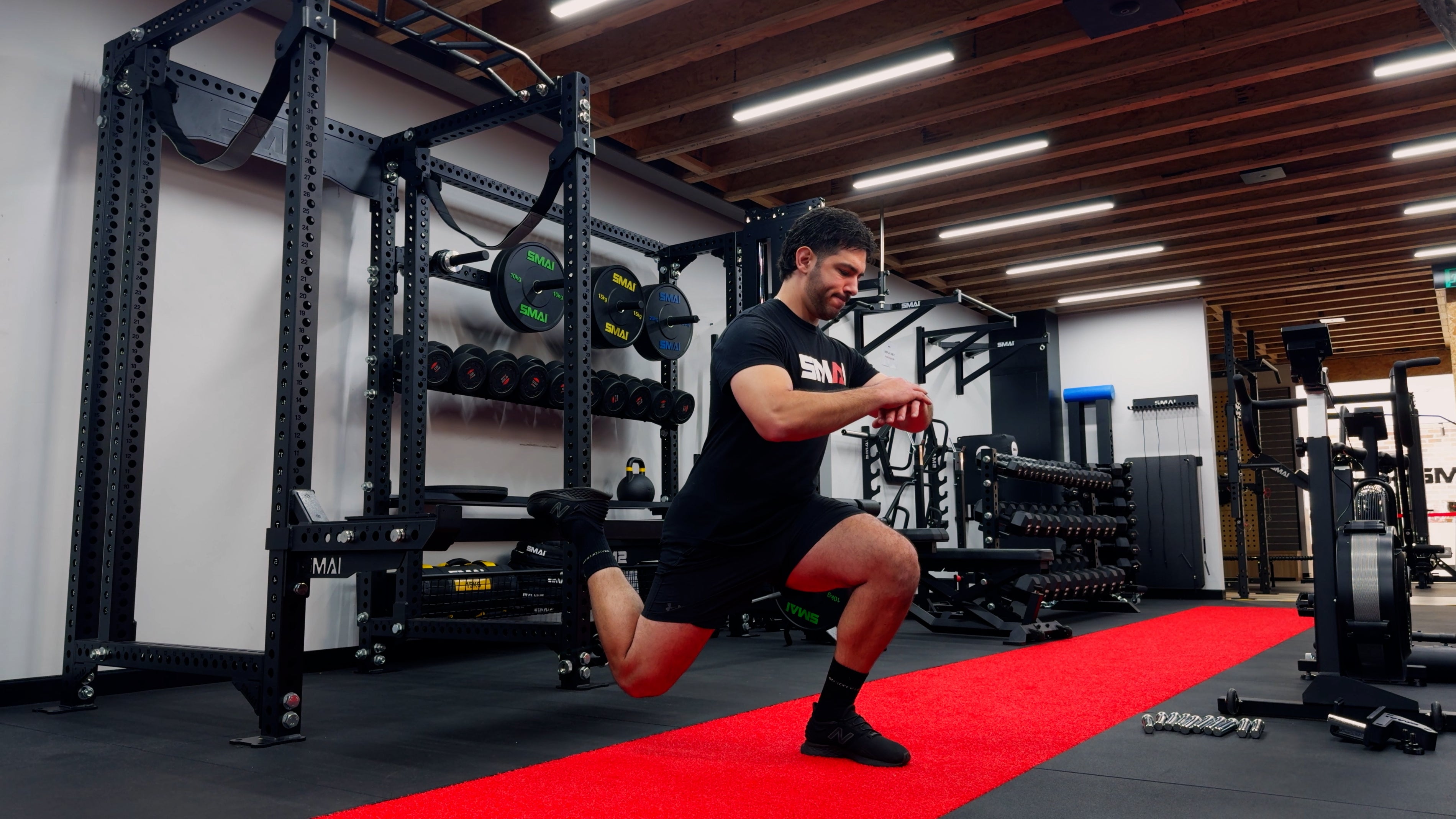 A man in athletic wear performs a Bulgarian split squat on a red mat in a gym, with one foot elevated on a bench behind him and weights and gym equipment in the background.