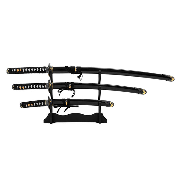 The SMAI Daisho - Carbon 3pc Set with Stand - Shi no Tora features three traditional Japanese swords with black and gold handles, displayed horizontally on a black wooden stand against a white background.
