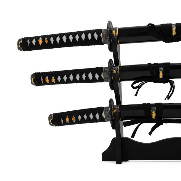 The SMAI Daisho - Carbon 3pc Set with Stand - Shi no Tora features three collectible samurai swords with black handles and gold accents, elegantly displayed on a black wooden stand against a white background.