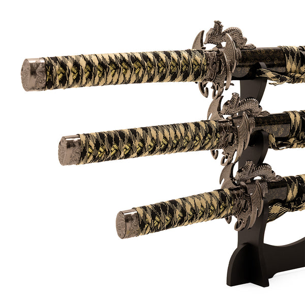 The SMAI Daisho 3pc Set with Stand - Desert Dragon features three elegant Japanese display swords with gold and black handles and dragon-themed guards, arranged horizontally on a black stand for striking decoration.