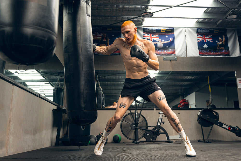 A shirtless man with orange hair and tattoos punches a heavy bag in a gym. He wears black shorts, white boxing shoes, and gloves. Australian flags and gym equipment are visible in the background.
