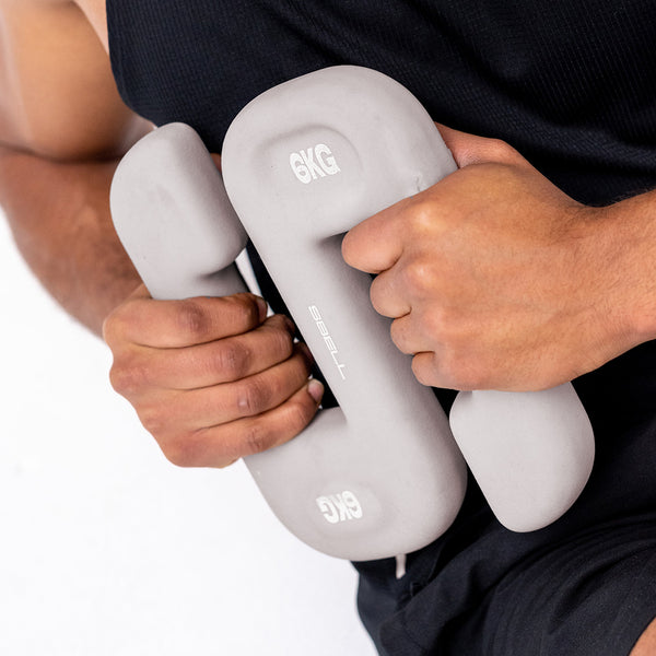 A person in a black tank top holds a pair of SBELL Grey 6kg dumbbells close to their chest. The focus is on the SBELL dumbbells, ideal for free weight training, and each one is labeled with "6KG" and "TPB06.