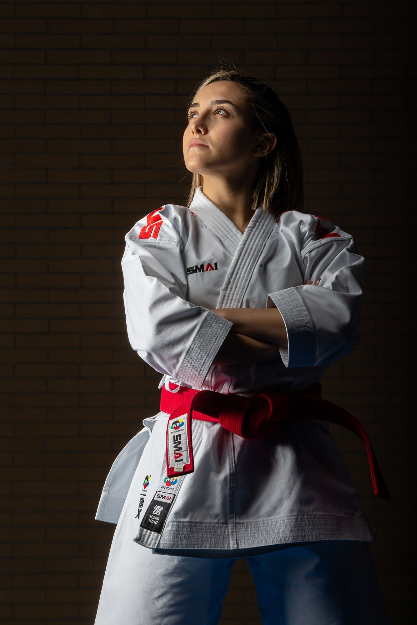 A woman in a karate gi with a red belt stands confidently with arms crossed, looking upward. The background is dark with a brick pattern, and she is lit dramatically from the front.