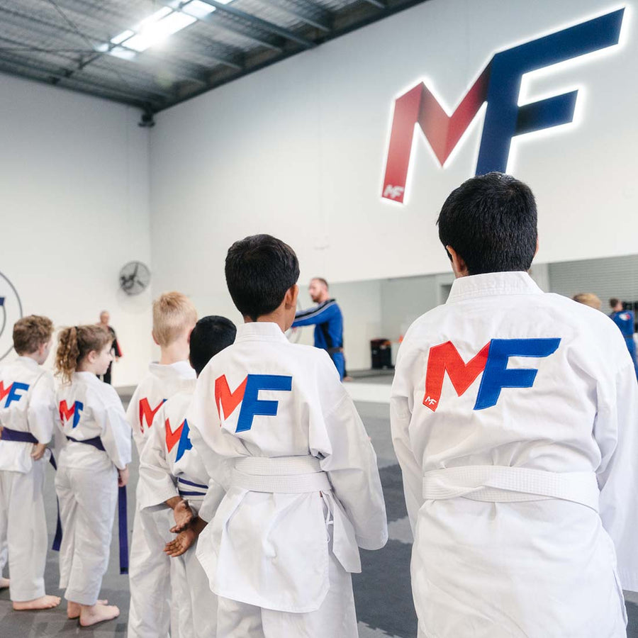 A group of children wearing white martial arts uniforms with "MF" logos on their backs stand in a line facing an instructor in a dojo setting. The room is spacious with a high ceiling and natural light.