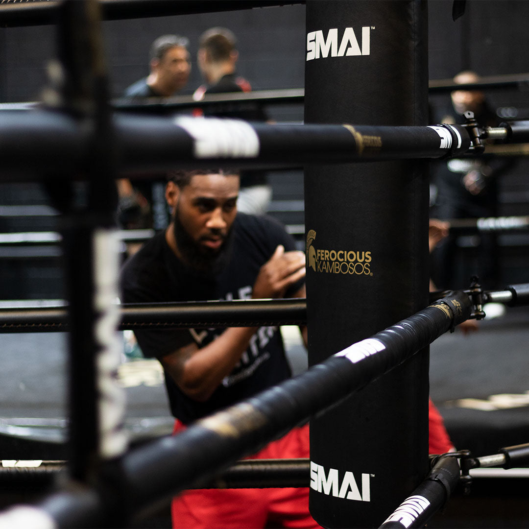 A man in a black shirt and red shorts trains in a boxing ring, throwing a punch. The image is partially obscured by the ropes of the ring, with a padded post labeled "Ferocious Kambosos" in the foreground.