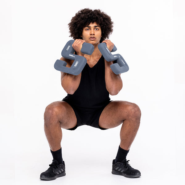 A person with curly hair is performing a squat exercise, demonstrating strength and conditioning, while holding two SBELL Midnight 12kg dumbbells from No Sync To Channel Engine. They are wearing a black tank top, black shorts, and black sneakers in a commercial gym setting against a white background.