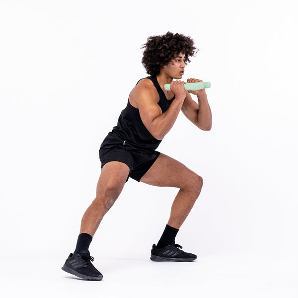 A person with curly hair, dressed in a black tank top and shorts, performs a squat while holding the SBELL Advanced pack dumbbell as part of strength and conditioning training. They are wearing black sneakers and standing against a plain white background.