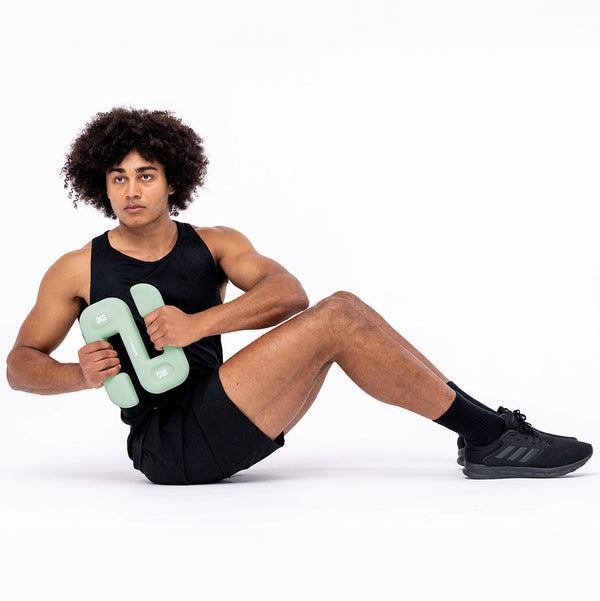 A person with curly hair, wearing a black tank top and shorts, demonstrates free weight training using SBELL Mint 8kg weights in a seated exercise. Set against a white background, the individual completes their workout outfit with black sneakers.