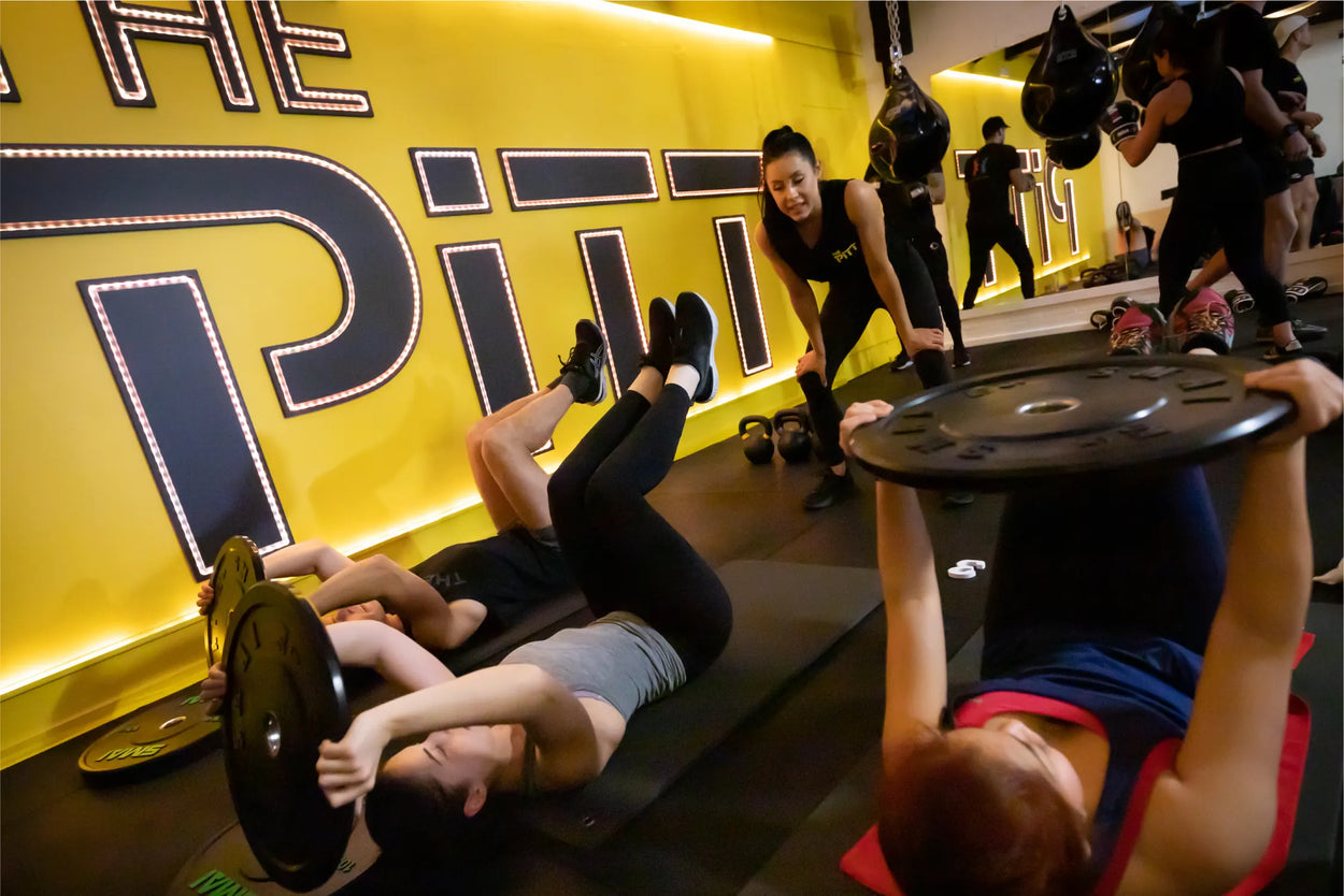 A group of people exercise on mats in a gym, lifting weights as an instructor in black observes and encourages them. The bright yellow wall behind them features large illuminated letters spelling "PITT.