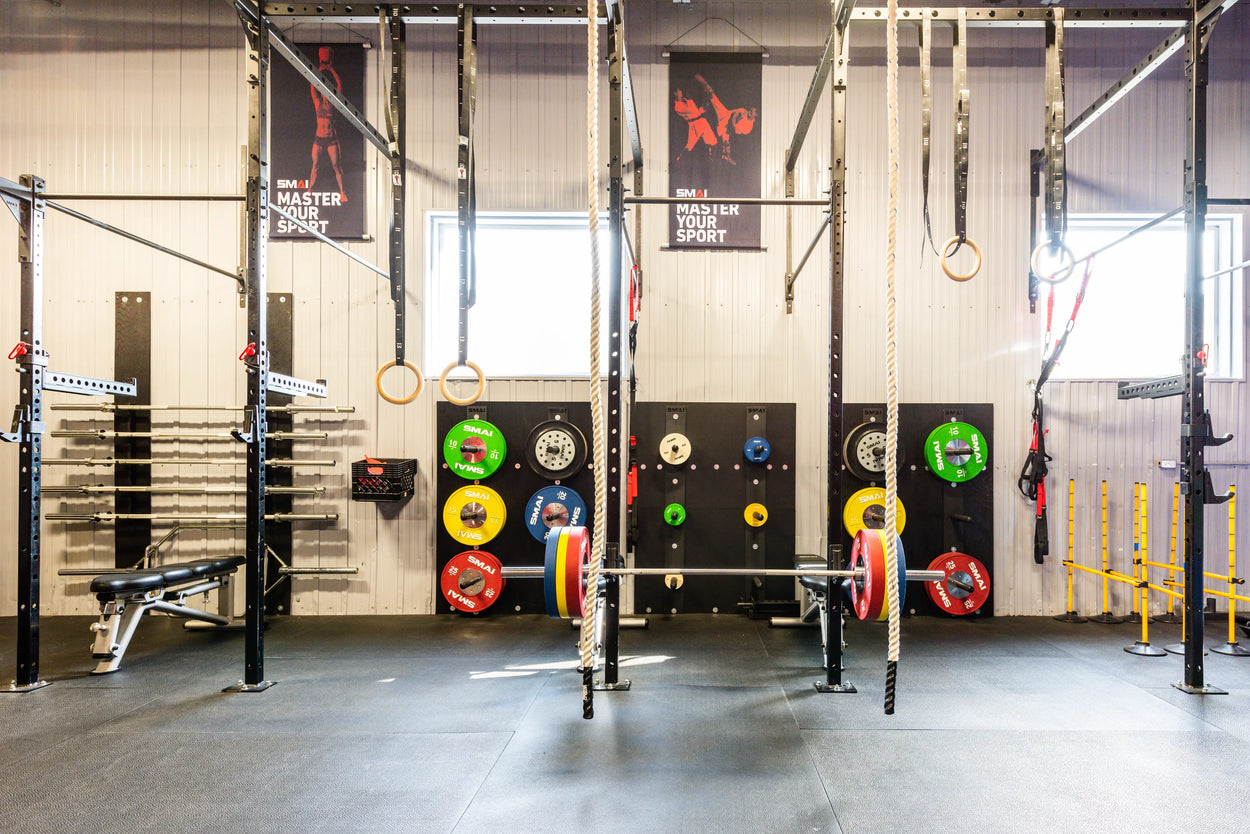 A gym with a variety of equipment, including barbells, weight plates in vibrant colors, gymnastic rings, ropes, and a bench. Posters on the wall say "Master Your Sport." The room is well-lit with natural light from two windows.
