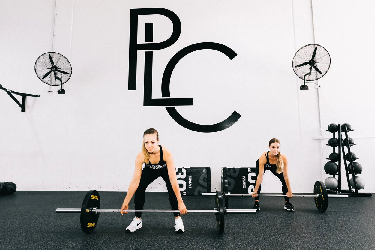 Two women in athletic wear perform deadlifts with barbells at a gym. A large "PLC" logo is on the wall behind them, along with two fans and stacked weights.