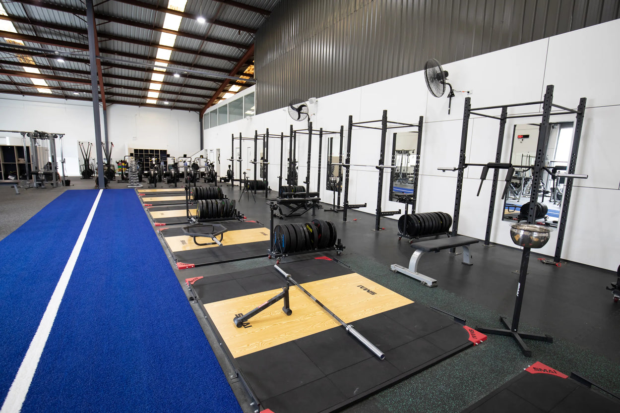 A spacious gym with weightlifting platforms, barbells, benches, and racks lined up next to a blue running track inside a high-ceilinged, well-lit facility.