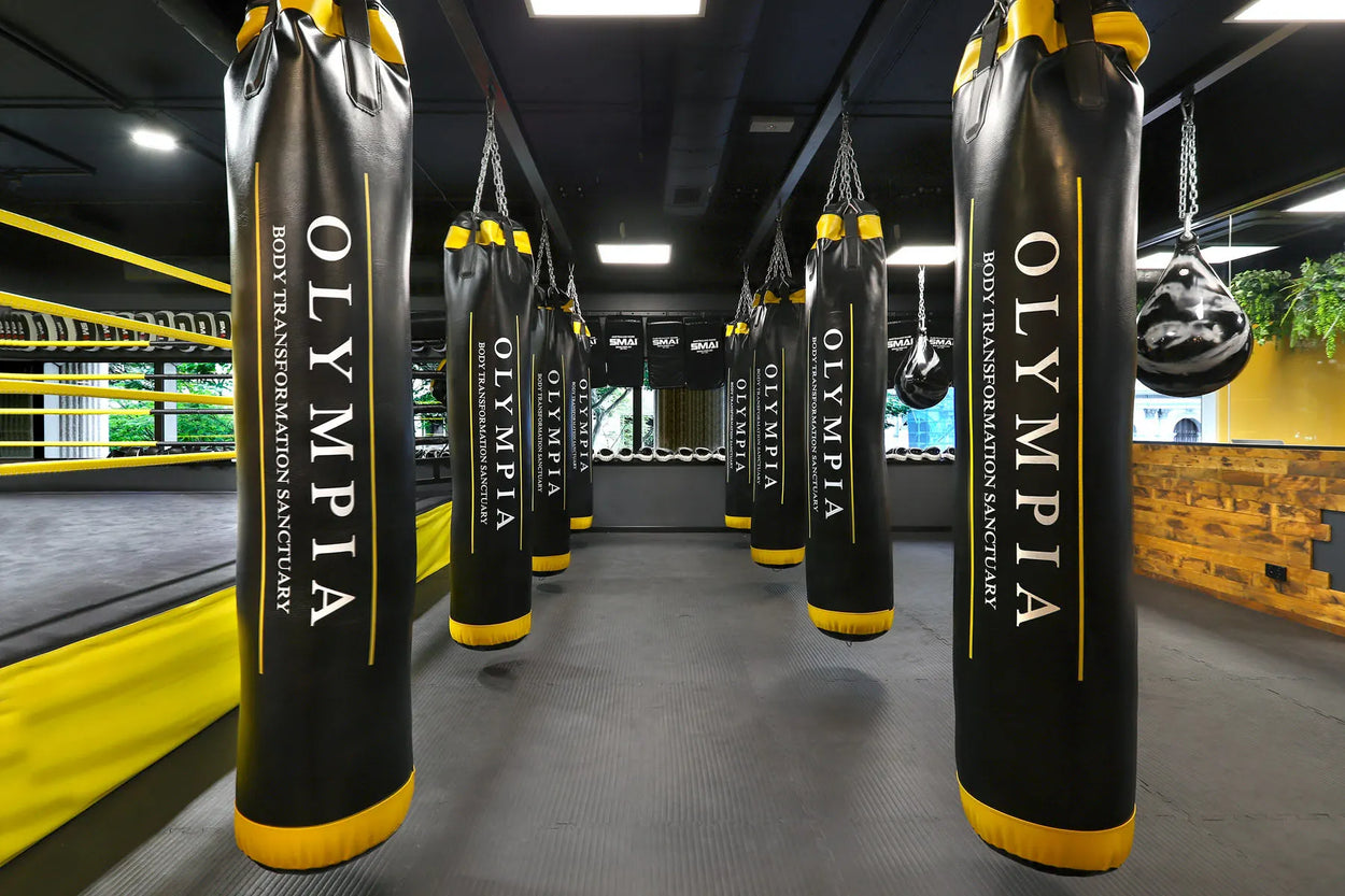 A modern boxing gym with multiple black and yellow punching bags labeled "Olympia" and "Body Transformation Sanctuary," a boxing ring on the left, and a wooden feature wall on the right.
