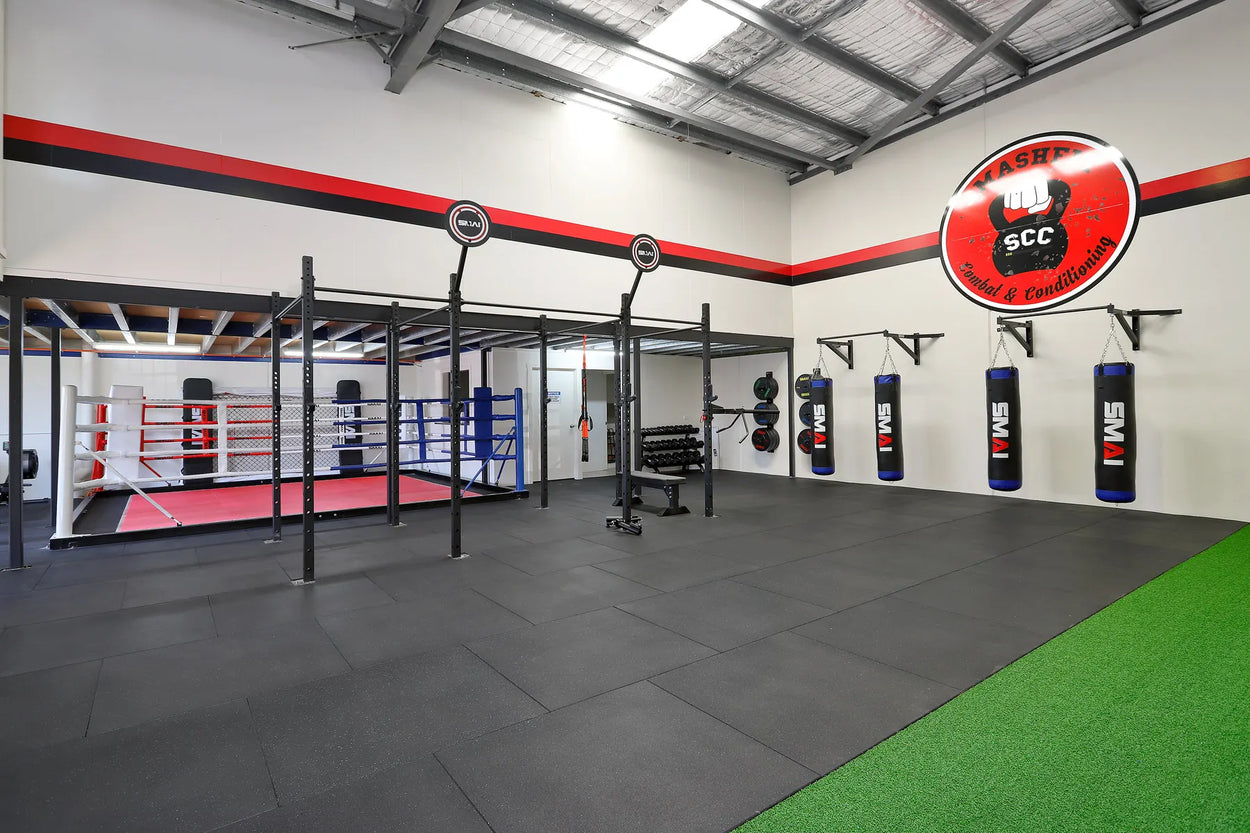 A spacious gym featuring a boxing ring, punching bags, gymnastic rings, and weightlifting equipment. The interior has a high ceiling and industrial design, with a logo on the wall and a green turf area on the right.