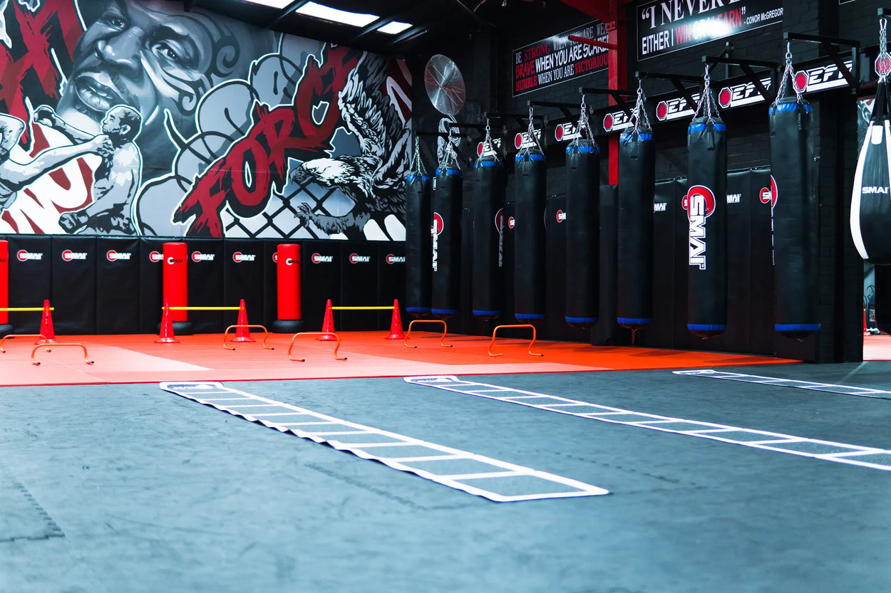 A spacious gym with red and black flooring, punching bags hanging in a row, agility ladders on the floor, and graffiti art on the walls featuring a large word "FORGE" and a boxer.