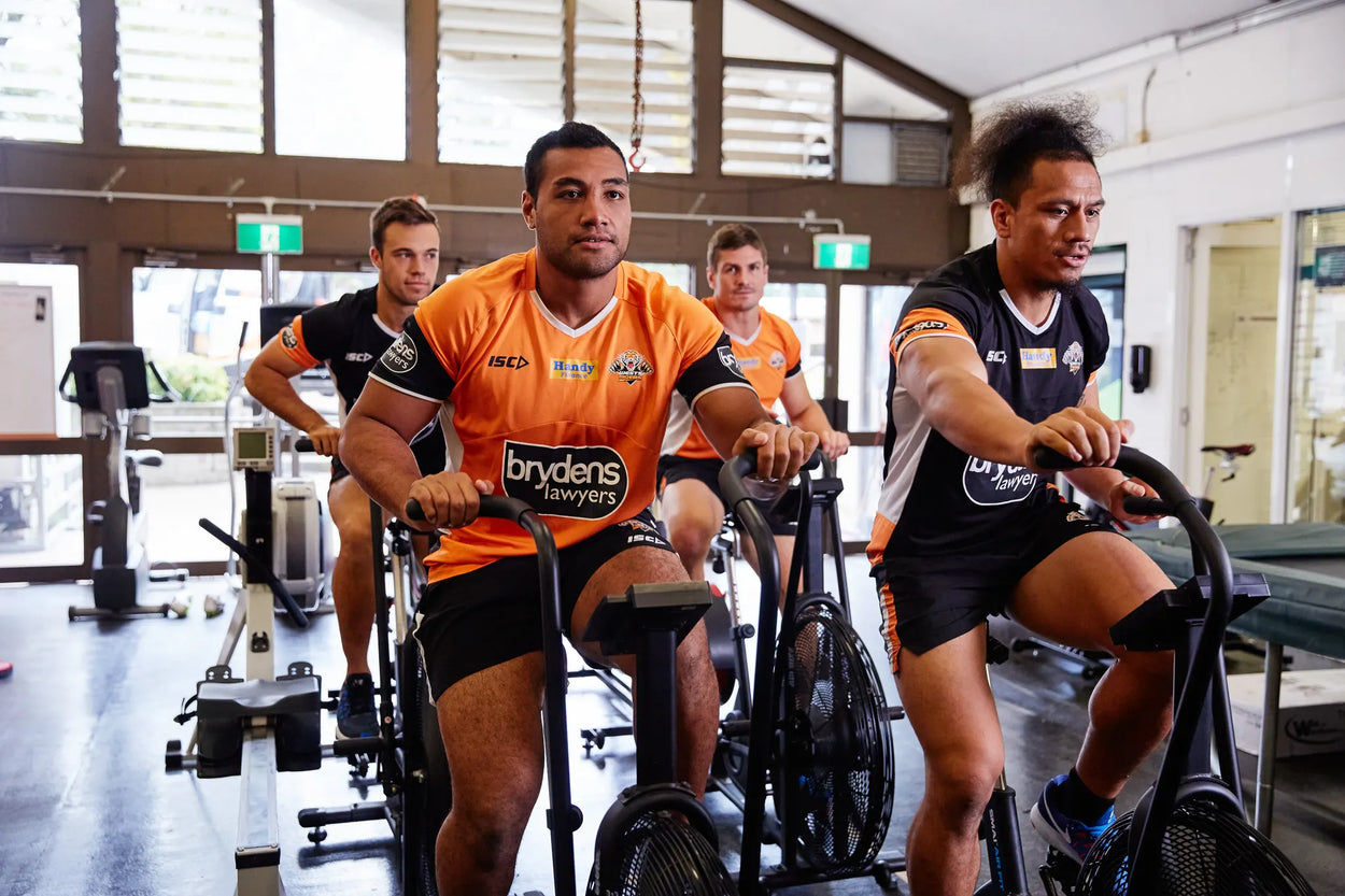 Four men wearing sports attire are riding stationary bikes in a gym. Two are in orange jerseys and two are in black. They appear focused and engaged in their workout. Gym equipment and large windows are visible in the background.