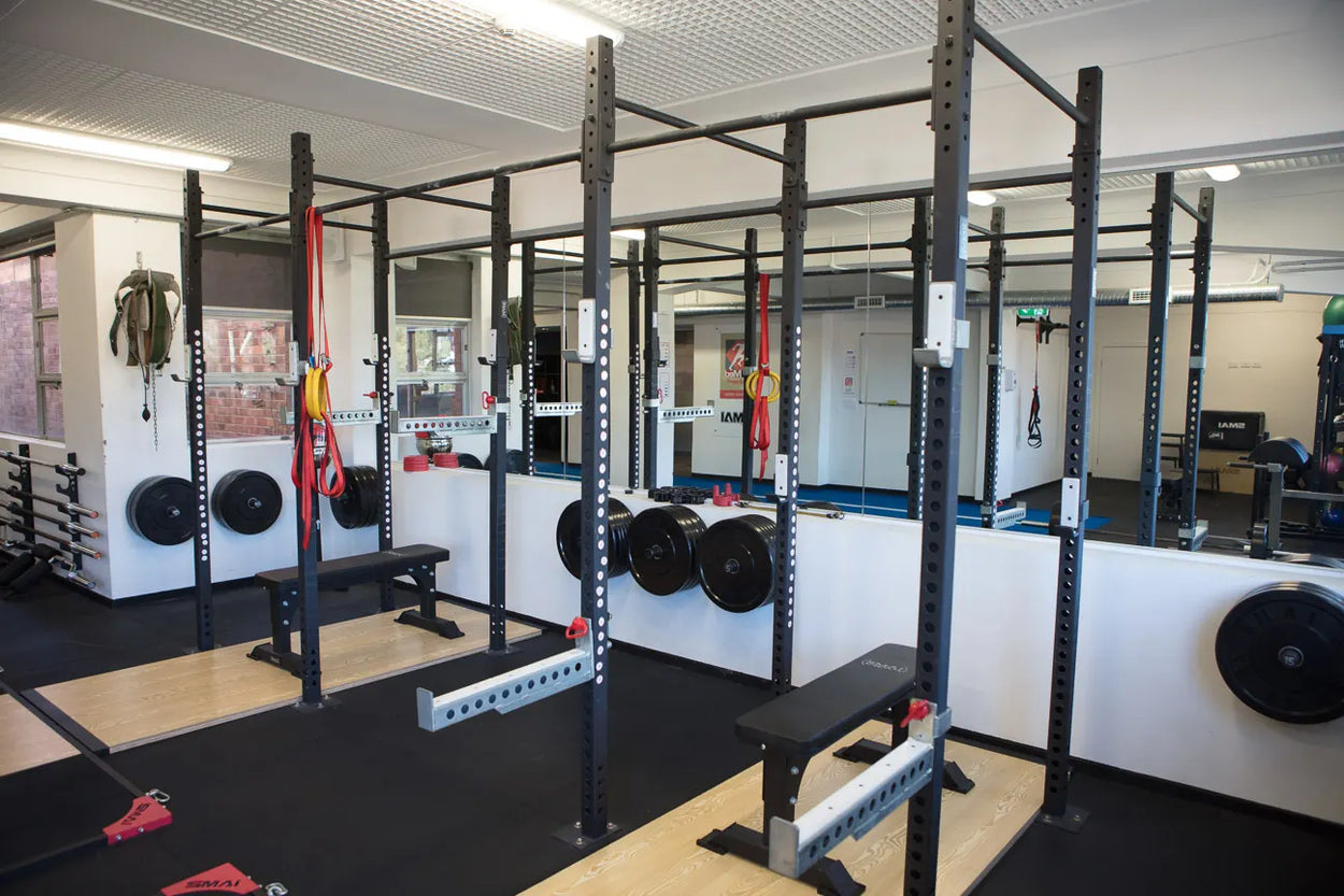 A gym with weight racks, benches, and various exercise equipment. The room is bright, with mirrors on the wall and a black rubber floor. Suspended bands and weight plates are visible throughout the space.