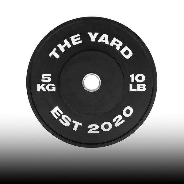 A black weight plate with white text reading "THE YARD," "5 KG," "10 LB," and "EST 2020" around the center hole.
