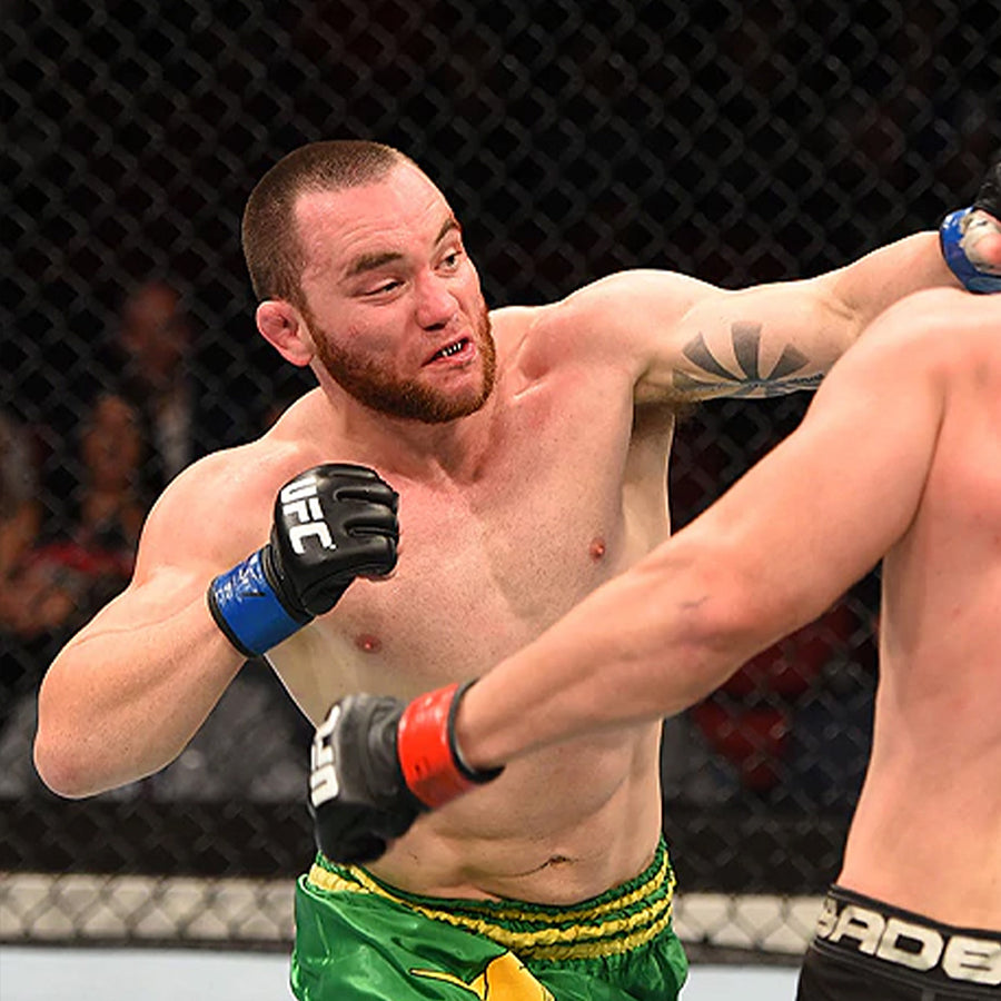 A mixed martial artist in green and yellow shorts throws a punch at his opponent inside an octagon cage during a UFC fight.