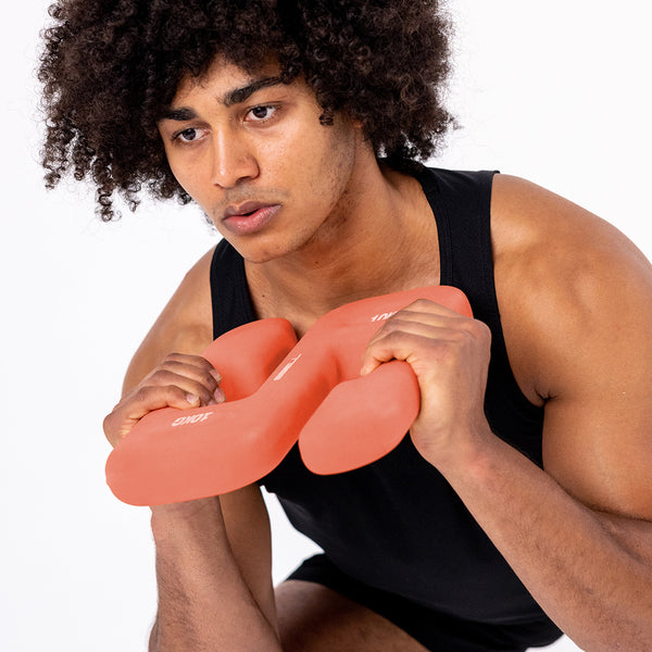 Wearing a black tank top, a person with curly hair holds an SBELL Advanced pack dumbbell close to their chest, capturing the essence of free weight training against a plain white backdrop.
