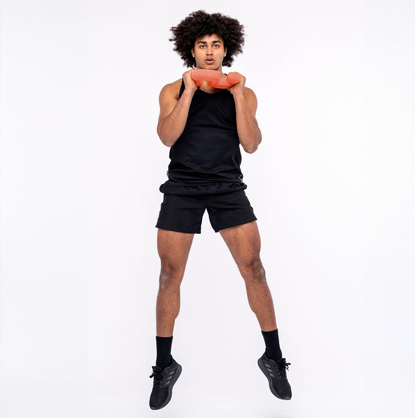 A person dressed in athletic gear, wearing a black tank top and shorts, performs a jump while holding an SBELL Advanced pack orange weighted disc at chest level, showcasing their strength and conditioning training. They are positioned against a plain white background.