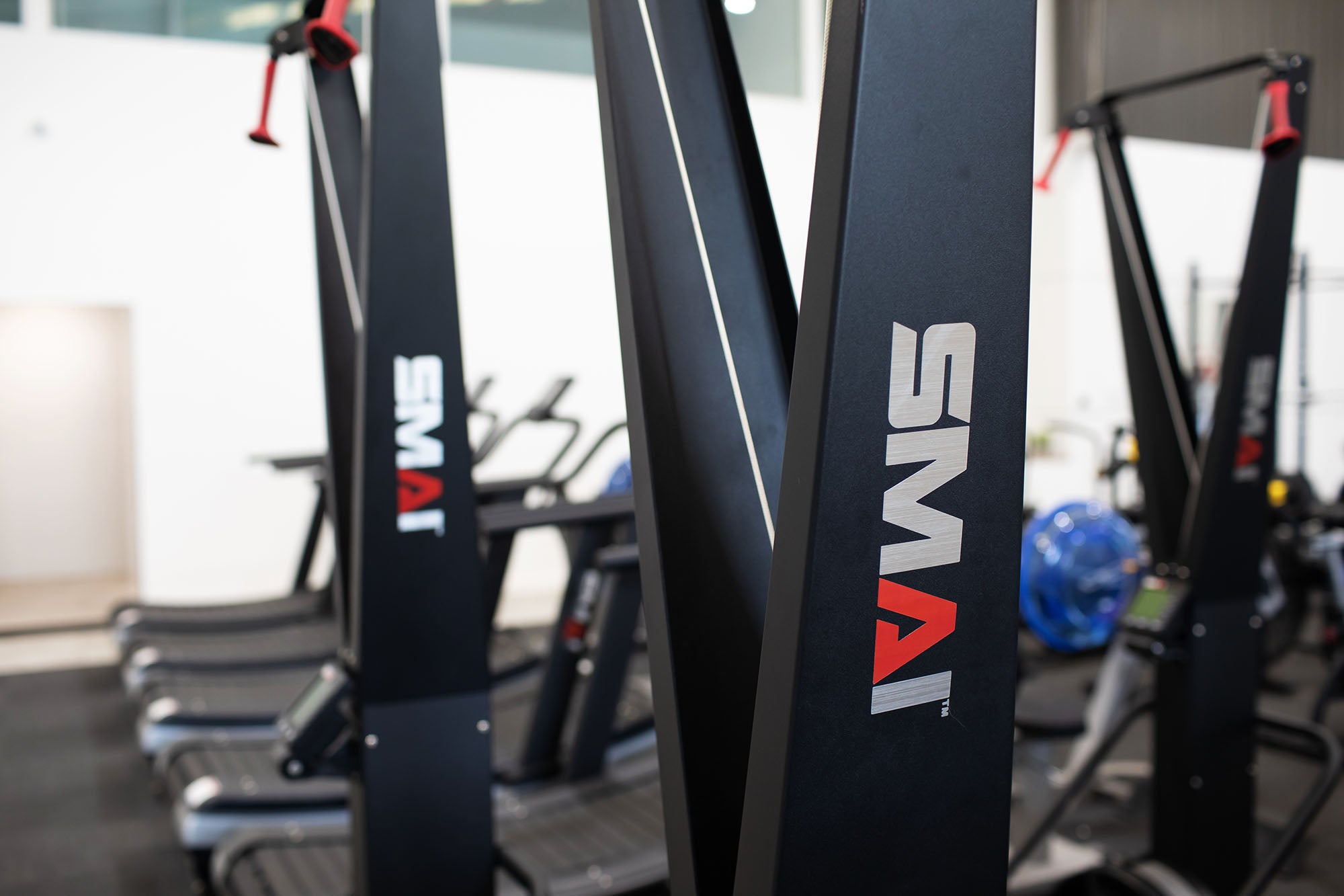Close-up of black gym equipment with the brand name "SMAI" in white and red lettering, with other fitness machines and treadmills visible in the blurred background.