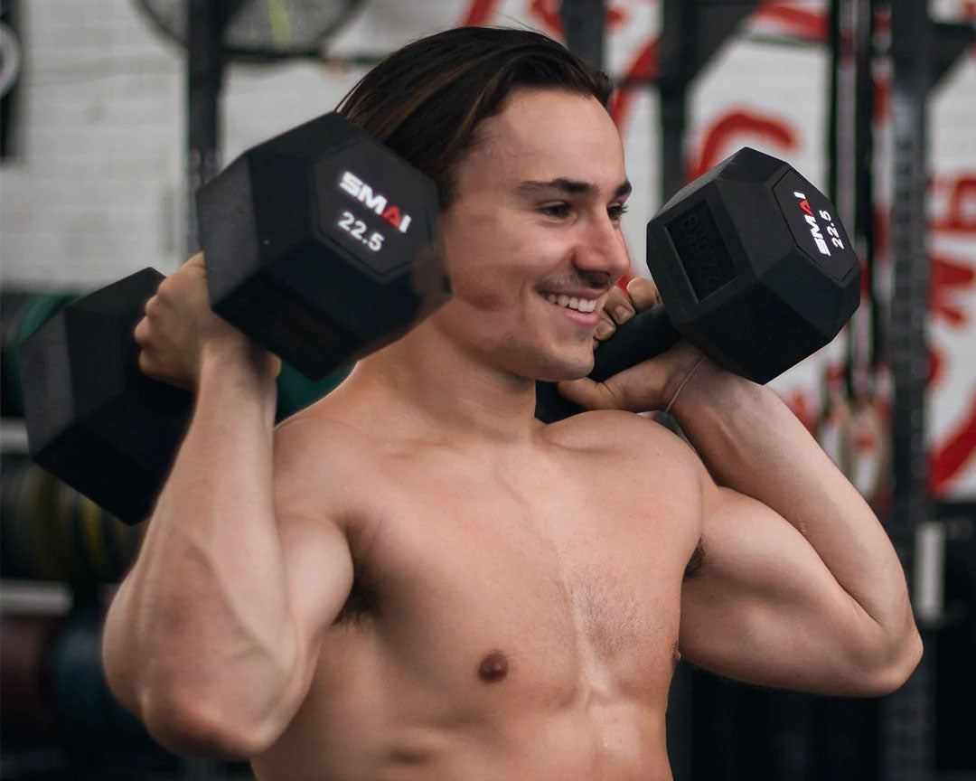 A shirtless man smiles while holding two black 22.5 kg dumbbells on his shoulders in a gym, with gym equipment visible in the background.