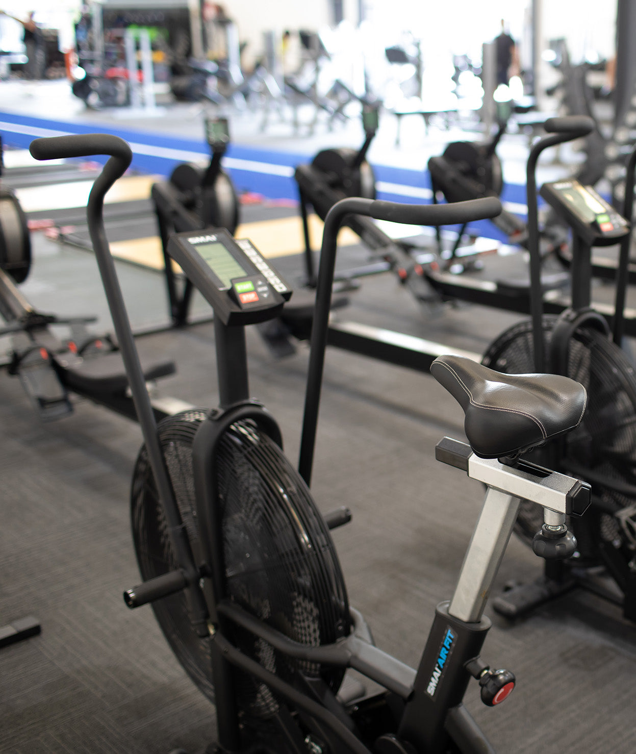 A row of stationary exercise bikes in a gym. The bikes have digital displays and are positioned next to blue mats and rowing machines. The gym has a bright, spacious atmosphere.