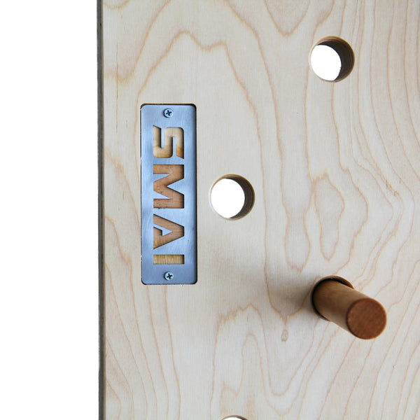Close-up of the SMAI Monster Peg Board: light marine ply with round holes, a metal "SMAI" plate, and a wooden peg inserted—perfect for load-bearing wall mount setups and intense peg board workouts.