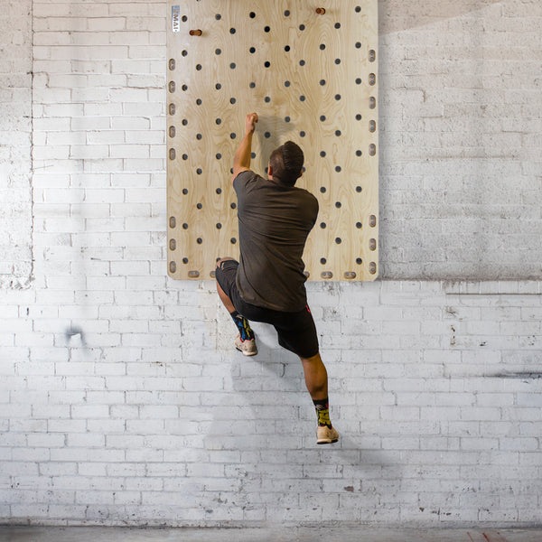 Wearing athletic clothing, a person climbs the SMAI Monster Peg Board, made of marine ply and fixed to a white brick wall, gripping pegs with their hands and supporting their weight with their legs.