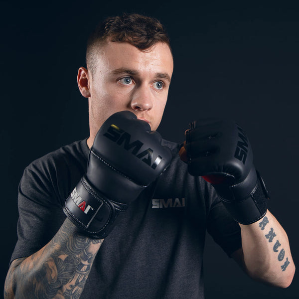 A man with tattoos, dressed in a dark SMAI shirt, poses in a fighting stance against a dark background while wearing SMAI Elite85 MMA Gloves in black.