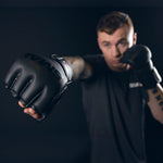 A person wearing SMAI Elite85 MMA Gloves in black, crafted from durable cowhide leather, throws a punch toward the camera with their other fist raised defensively against a dark background.