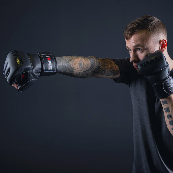 A man with tattoos, wearing a dark shirt and SMAI Elite85 MMA Gloves in black, throws a punch against a dark background.