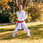 A person in a white SMAI WKF Reversible Karate Uniform - 6oz Premium Kumite Gi - Inazuma RX with a red belt stands barefoot on grass in a martial arts pose, surrounded by trees and sunlight.