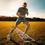 A tattooed man in athletic wear and white sneakers uses the SMAI Rubber Rollout Agility Ladder for speed drills on a grassy field at sunset.