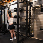 A person in a gym reaches for a pull-up bar on a black workout rack, with the SMAI Cable Pulley System nearby. They wear a white tank top, black shorts, and white sneakers, surrounded by functional strength training equipment.