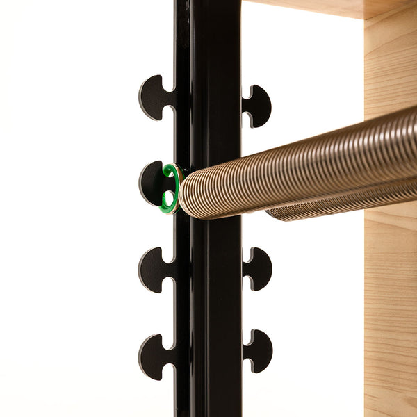 Close-up of a metal spring with curved hooks attached to the black frame of the SMAI Pilates Chair, next to a wooden platform—perfect for core conditioning and strength training—set against a white background.