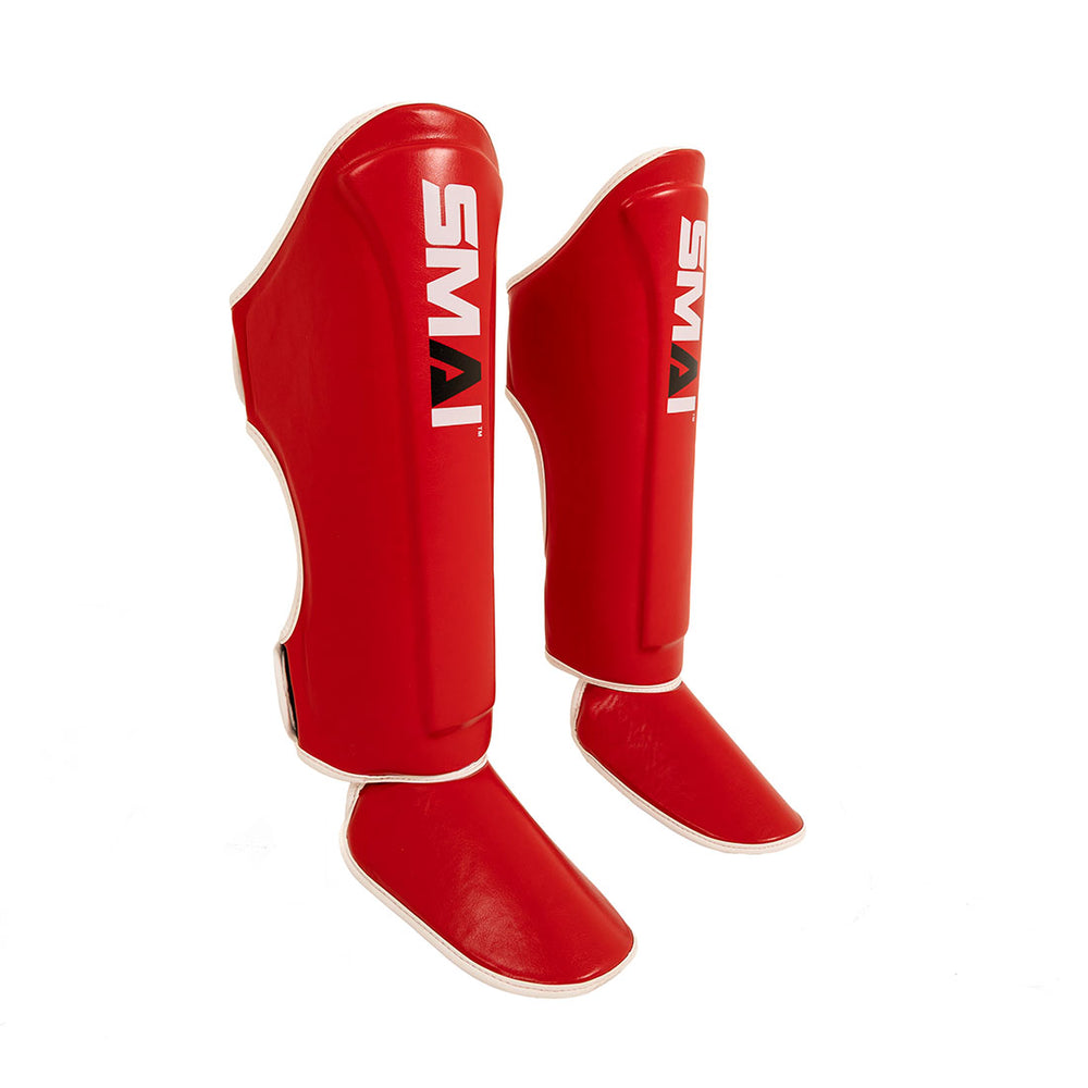 The SMAI Essentials Muay Thai Shin Instep Guard - Red features synthetic leather, attached foot protectors, and a white SMAI logo. The pair is shown upright on a white background.