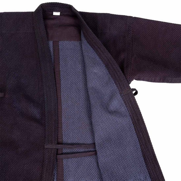 A close-up of the SMAI Deluxe Kendo Jacket - Indigo Double Layer Keikogi with the front open, revealing its textured inner lining and lapels. The right sleeve, made from 100% cotton, is extended outward.