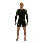 A man stands barefoot on a white background, wearing a black and olive green long-sleeve rash guard and the SMAI MMA Grappling Shorts - Black / Olive Green, both featuring visible "SMAI" logos. The shorts include a stretch fabric gusset for enhanced mobility and comfort.