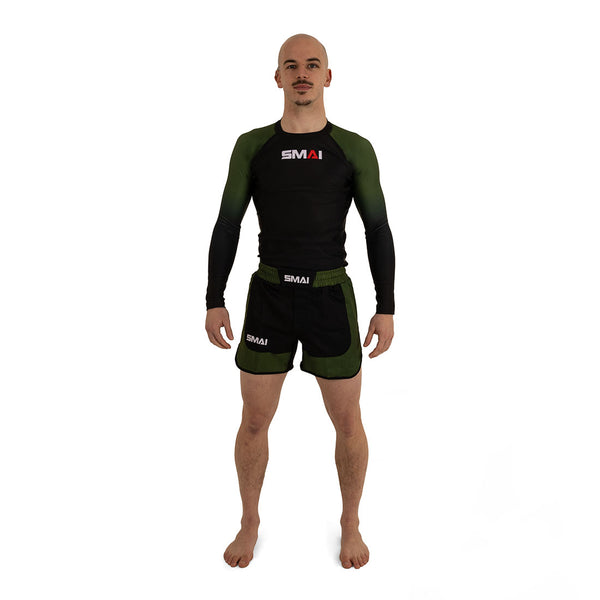 A man stands barefoot on a white background, wearing a black and olive green long-sleeve rash guard and the SMAI MMA Grappling Shorts - Black / Olive Green, both featuring visible "SMAI" logos. The shorts include a stretch fabric gusset for enhanced mobility and comfort.
