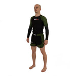A man stands barefoot against a plain white background, wearing SMAI MMA Grappling Shorts in Black and Olive Green, featuring a stretch fabric gusset.
