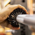 A hand uses the SMAI Barbell Cleaning Brush - Steel to scrub the knurling on a metal barbell in a gym setting.